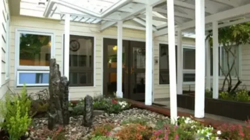 A front entrance with a covered walkway and white columns, surrounded by a landscaped garden with rocks and flowering plants