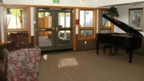 A small-scale image of an indoor common area with a floral armchair and a grand piano near glass doors that lead to an outdoor patio